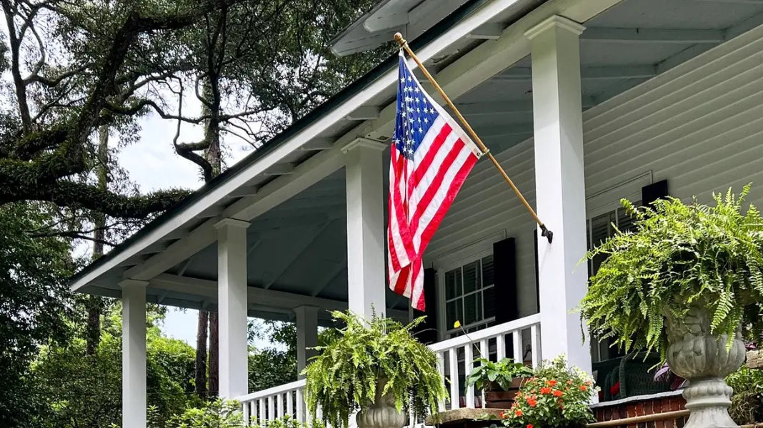 American-made flag hangs from angled wooden pole on a white porch adorned with ferns and red flowers.