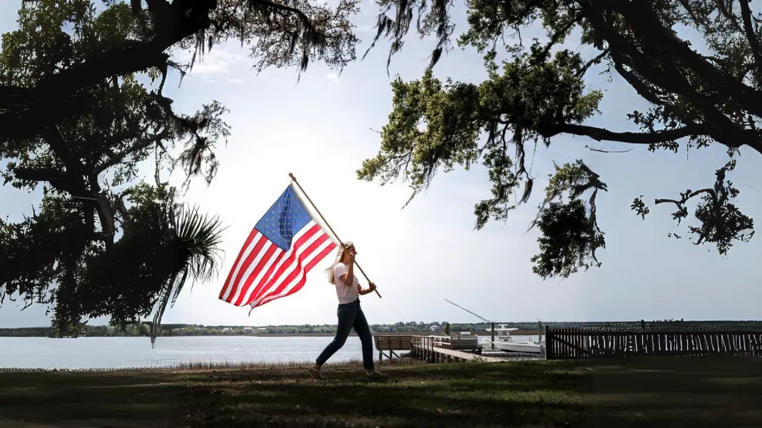 American Flag displayed as a large flag carried by a woman near a lakeside under oak trees.
