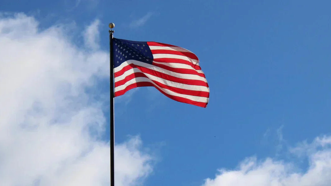 American flagpole with the U.S. flag waving high beside a historic building under a bright blue sky.
