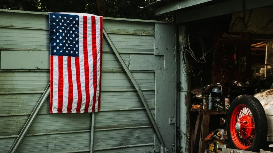 American flag hanging on a weathered storage shed beside an old garage with a vintage car wheel.