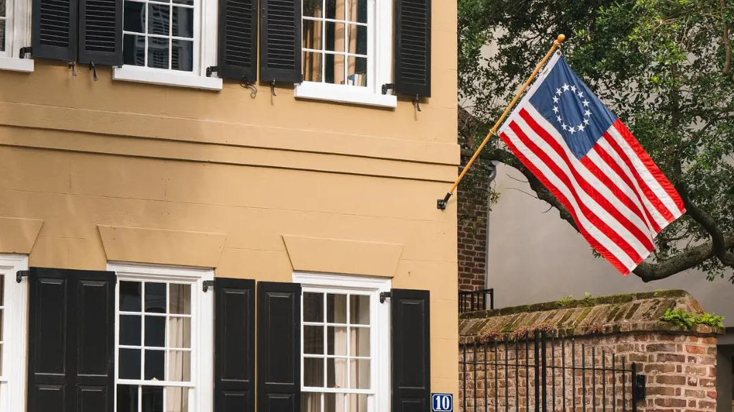Betsy Ross flag mounted on a tan building with black shutters, waving beside a historic brick courtyard