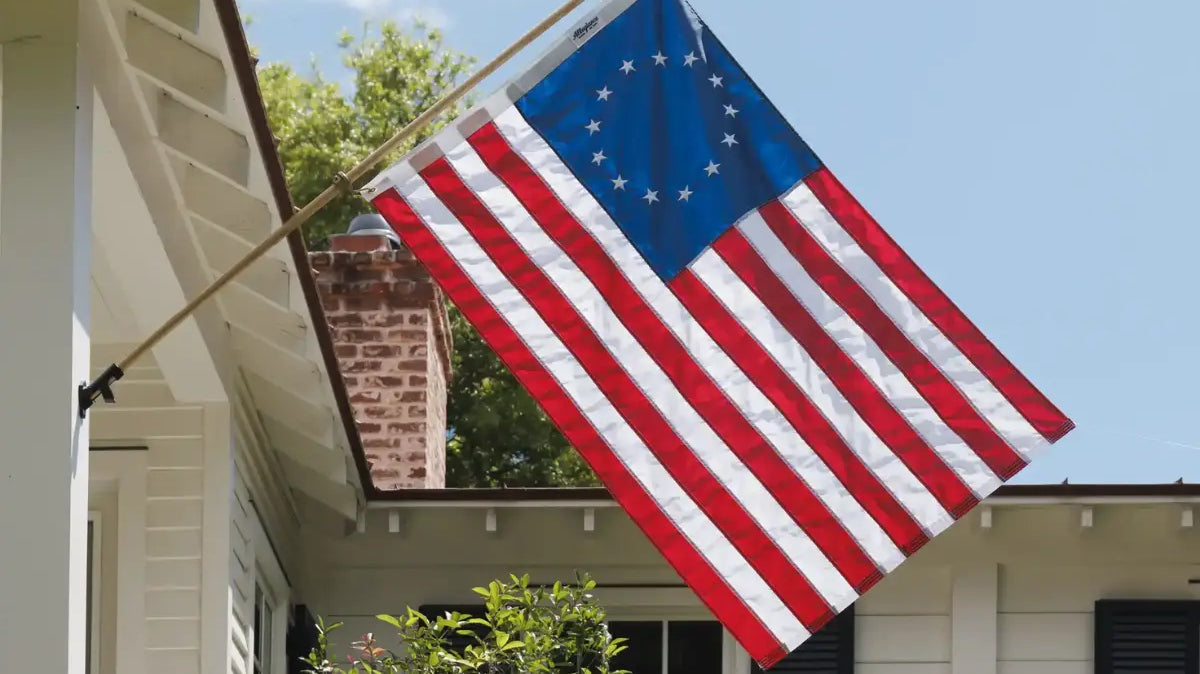 Betsy Ross flag mounted on a white house with a brick chimney and greenery in the background.