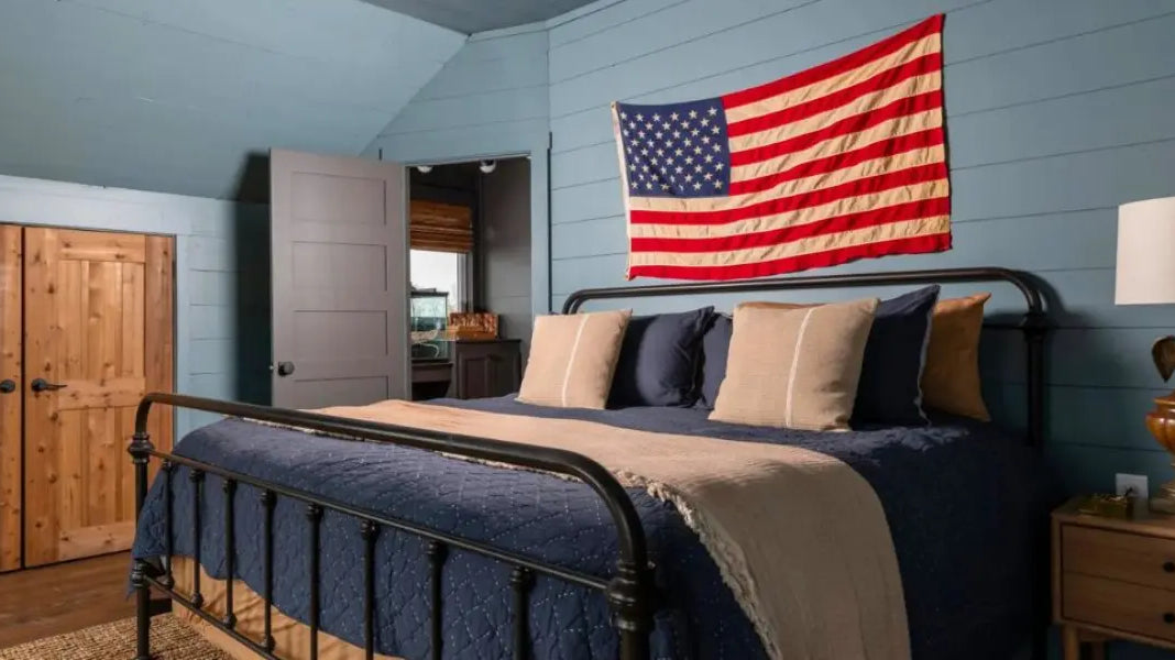 High-quality American flag hanging above a rustic metal bed in a cozy, blue-paneled bedroom.