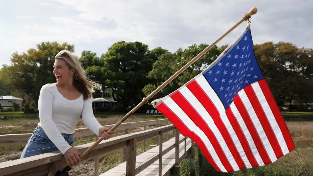 High-quality U.S. flag waving as a smiling woman holds it on a wooden dock with greenery and houses in the background.