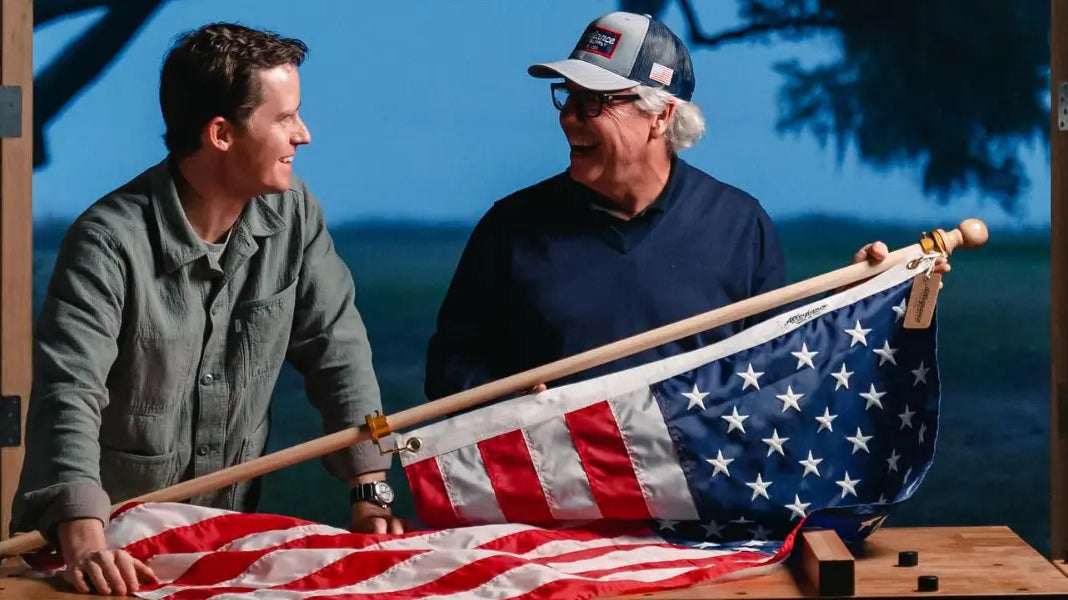 Patriotic American flag held by two smiling men inside a wooden structure with a scenic outdoor background