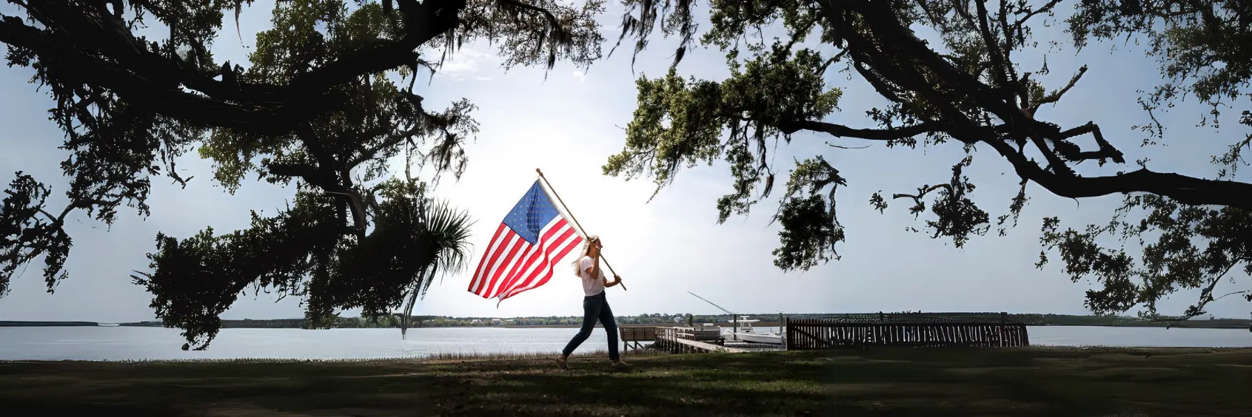 American Flag displayed as a large flag carried by a woman near a lakeside under oak trees.