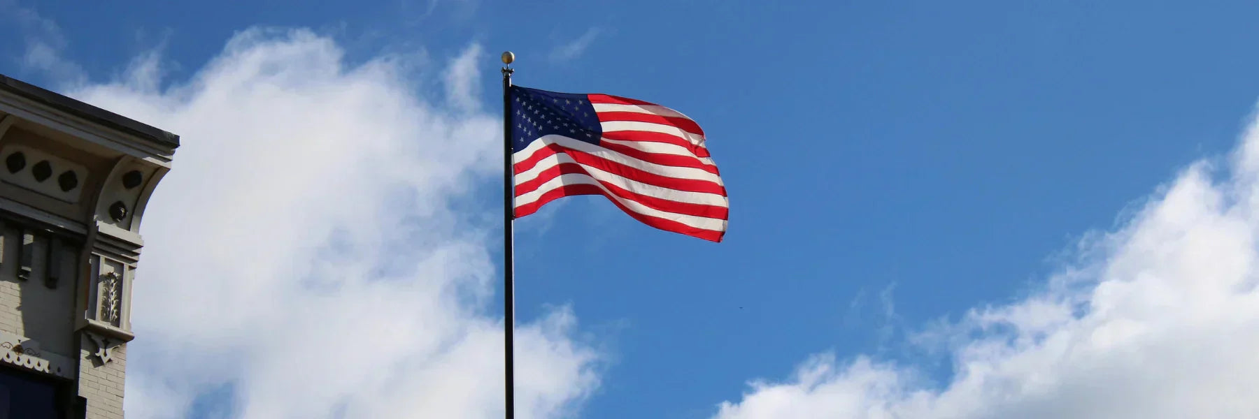 American flagpole with the U.S. flag waving high beside a historic building under a bright blue sky.