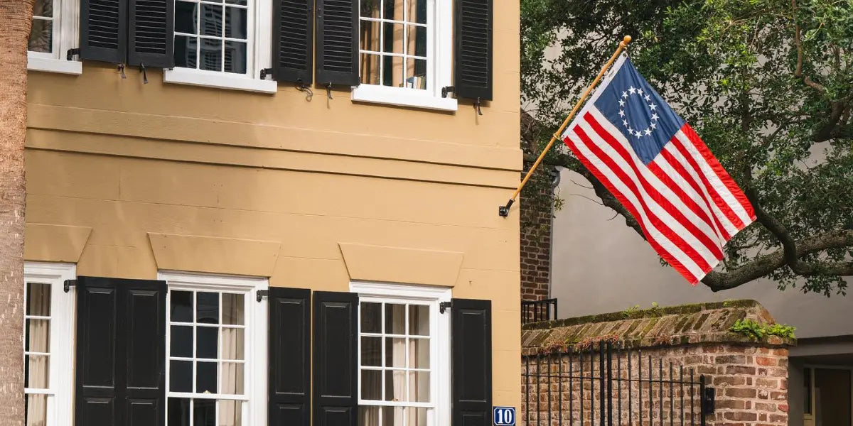 Betsy Ross flag mounted on a tan building with black shutters, waving beside a historic brick courtyard