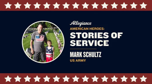 Mark Schultz stands with a young boy dressed in patriotic attire, surrounded by rows of flags in a grassy field