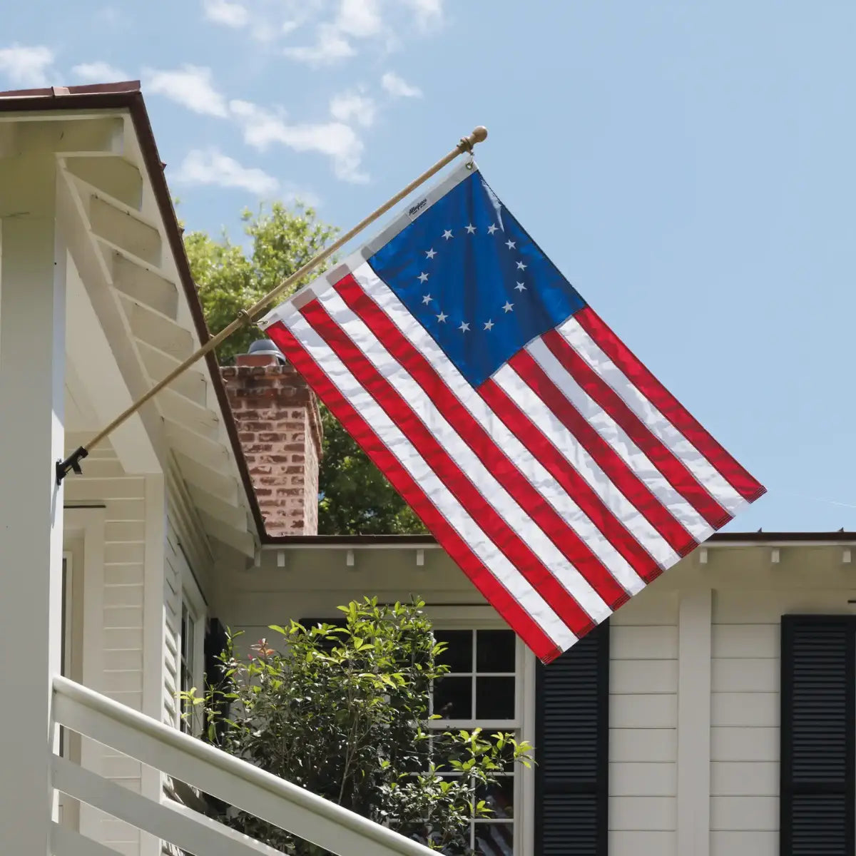 Betsy Ross flag mounted on a white house with a brick chimney and greenery in the background.